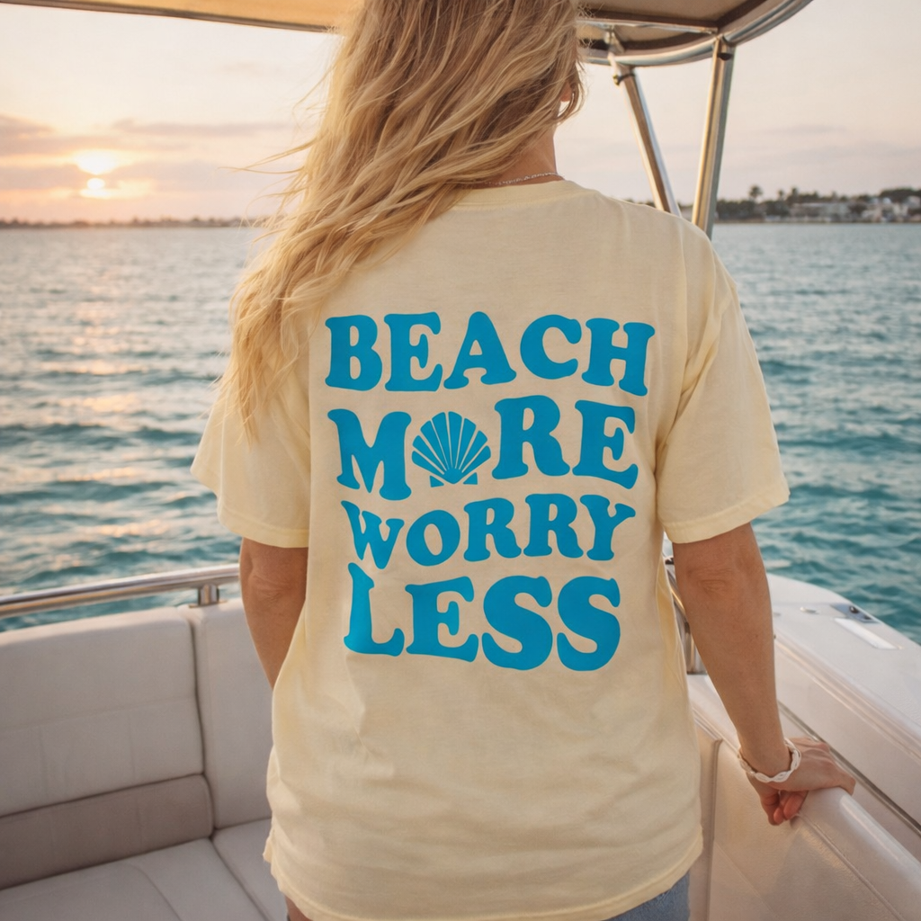 Yellow short sleeve t-shirt with blue text reading ‘Beach More Worry Less’ and a seashell graphic on the back.