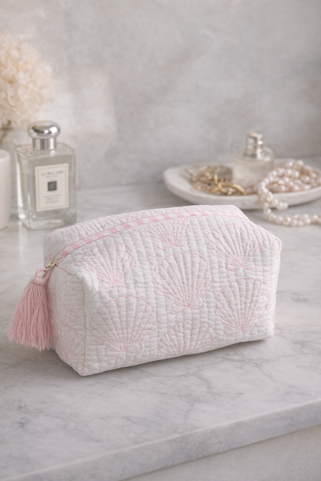 Light pink quilted cosmetic pouch with seashell pattern and tassel zipper on a marble surface.