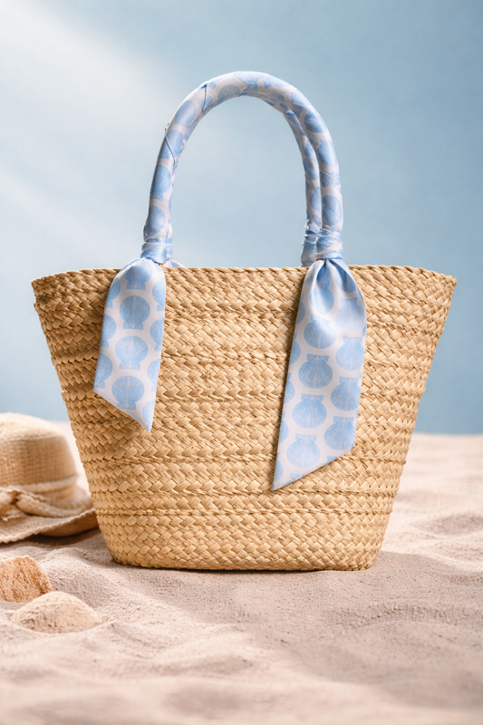 Woven straw bag with blue patterned handles on a sandy surface with a light blue background