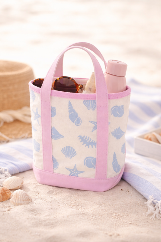 Small beach tote bag with pink trim and blue seashell pattern sitting on sand with empty interior.